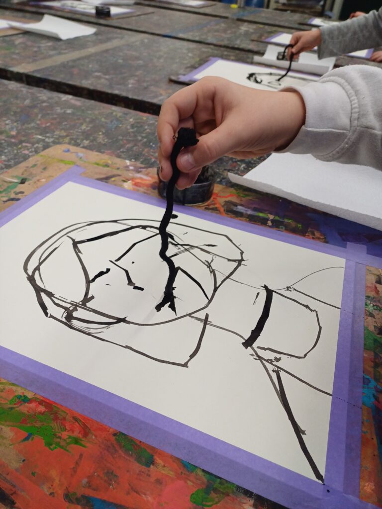 Kinder malen Gesichter mit schwarzer Farbe auf ein Stück Papier, das auf einen bunten Kunstschultisch geklebt ist